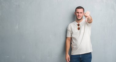 Beyaz delikanlı mutsuz ve kızgın ret gösterilen ve başparmak aşağı hareketi ile negatif seyir güneş gözlüğü takıyor gri grunge duvarın üzerinden. Kötü ifade.