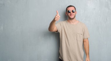 Gösterilen ve parmak gülümseyen süre bir numara ile kendinden emin ve mutlu yukarıyı güneş gözlüğü takıyor gri grunge duvarın üzerinden beyaz delikanlı.