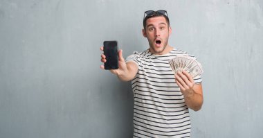 Dolar ve smarphone korkmuş şok bir sürpriz yüz, korkuyor ve heyecanlı korku ifadesi ile tutarak gri grunge duvarın üzerinden beyaz delikanlı