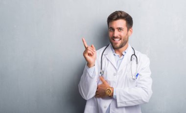 Yakışıklı genç adam yüzünde kocaman bir gülümseme ile gri grunge duvarın üzerinden kameraya bakarak yan el ve parmak ile işaret eden doktor.