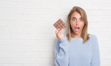 Beyaz tuğla duvar yeme üzerinde güzel genç kadın çikolata bar bir sürpriz yüz, korkuyor ve heyecanlı korku ifadesi ile şok korkmuş