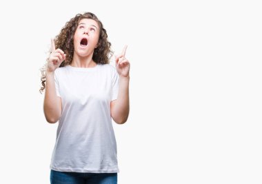 Güzel esmer kıvırcık saçlı Genç kız izole arka plan üzerinde rahat t-shirt giyen hayret ve yukarıya bakıp parmakları ile işaret şaşırttı ve silah kaldırdı.