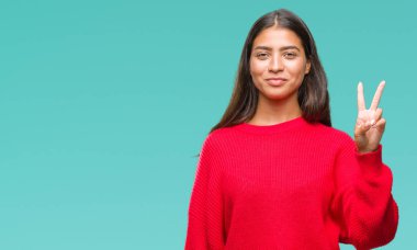 Genç güzel Arap kadın giyiyor kış kazak gösterilen ve parmakları ile işaret izole arka plan üzerinde ikincisi ise kendinden emin ve mutlu gülümseyerek.