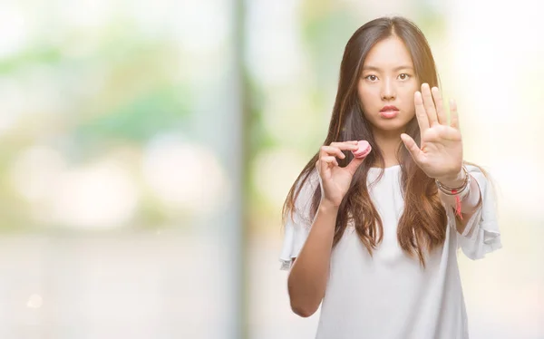 孤立の背景にピンクのマカロン甘いを食べる若いアジア女性は防衛ジェスチャー 深刻な自信を持って式に一時停止の標識をやって手を開いてください — ストック写真