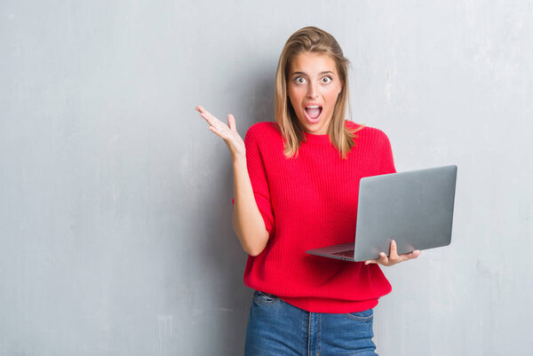 Beautiful young woman over grunge grey wall using computer laptop very happy and excited, winner expression celebrating victory screaming with big smile and raised hands