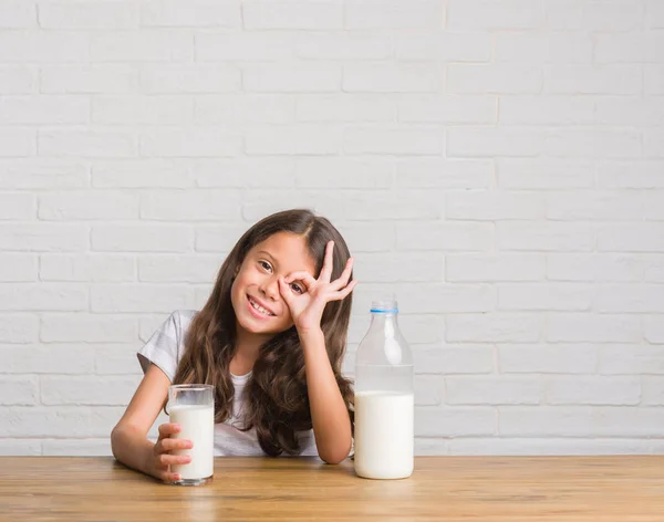 指を通して見る目に手で サインをして笑っている幸せそうな顔でミルクを飲んでテーブルに座って若いヒスパニックの子供 — ストック写真