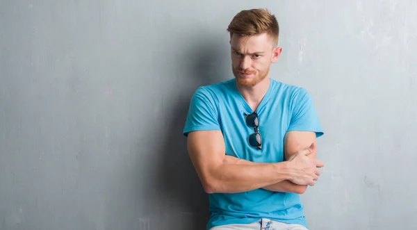 Young redhead man over grey grunge wall wearing casual outfit skeptic and nervous, disapproving expression on face with crossed arms. Negative person.