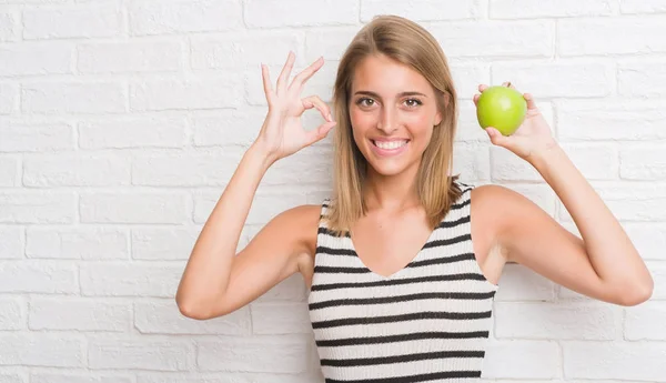 美丽的年轻女子在白色砖墙吃绿色苹果做确定的标志与手指 优秀的象征 — 图库照片