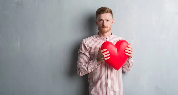 Young Redhead Man Grey Grunge Wall Holding Red Heart Confident — Stock Photo, Image