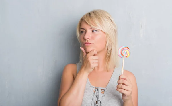 白种人成年妇女在灰色的壁上吃糖果棒棒糖严肃的脸思考问题 很困惑的想法 — 图库照片