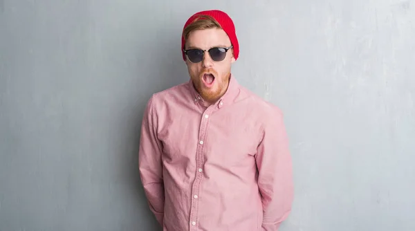 Young redhead man over grey grunge wall wearing wool cap and sunglasses scared in shock with a surprise face, afraid and excited with fear expression