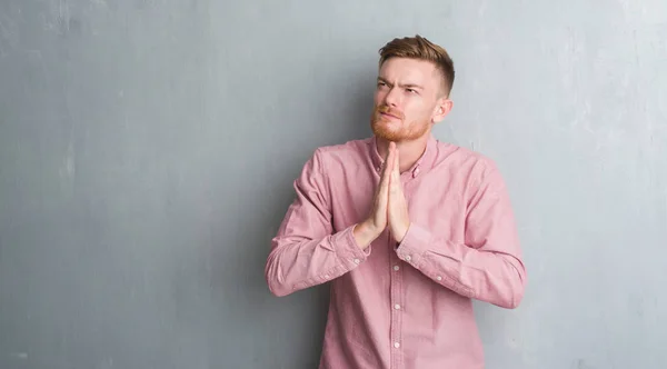 年轻的红发男子在灰色的格格墙上穿着粉色衬衫乞讨和祈祷 双手放在一起 希望表情在脸上非常情绪化和担忧 请求宽恕 宗教概念 — 图库照片