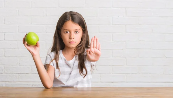 新鮮な青リンゴを食べるテーブルの上に座って若いヒスパニックの子供防衛ジェスチャー 深刻な自信を持って式に一時停止の標識をやって手を開く — ストック写真