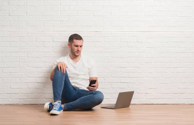 Acımak yüz ciddi düşünüyorum üstünde kendine güvenen bir ifade ile bilgisayar dizüstü ve smartphone kullanarak beyaz tuğla duvar üzerine oturan genç beyaz adam