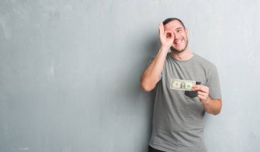 Mutlu yüz el parmaklarının arasından seyir göz üzerinde ok işaretiyle yapıyor gülümseyen bir dolar gösterilen gri grunge duvarın üzerinden beyaz delikanlı