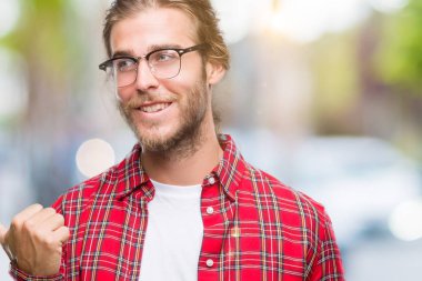 Genç yakışıklı adam uzun saçlı gözlüklü seyir ve başparmak yanında yukarıyı mutlu yüz ile gülümseyen izole arka plan üzerinde.