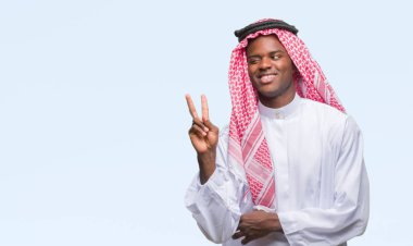 Mutlu yüz zafer işareti yaparken kameraya göz kırpıyor ile gülümseyen izole arka plan üzerinde geleneksel keffiyeh giyen genç Arapça Afrika adam. İki numaralı.