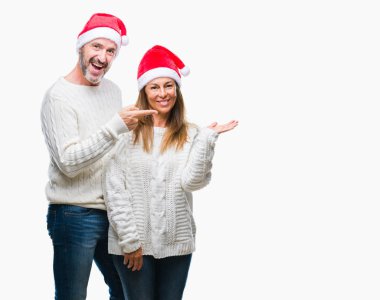 Orta yaş İspanyol çift hayrete ve el ile sunulması ve parmak ile işaret ederken kameraya gülümseyen izole arka plan üzerinde Noel şapka.