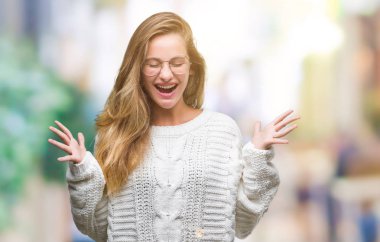 Genç güzel sarışın kadın giyiyor kış kazak ve kızgın ve silah ile başarı için deli kutluyor izole arka plan üzerinde güneş gözlüğü kaldırdı ve heyecanlı çığlık gözler kapalı. Kazanan kavramı