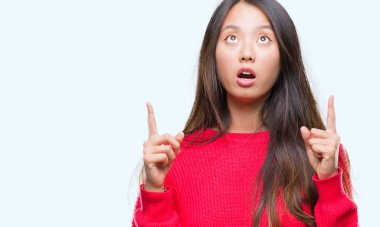 İzole arka plan üzerinde kış kazak giyen genç Asyalı kadın hayrete ve yukarıya bakıp parmakları ile işaret şaşırttı ve silah kaldırdı.