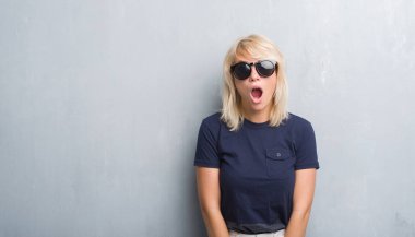 Grunge gri duvar korkuyor ve şok sürpriz ifade, korku ve heyecan yüz ile güneş gözlüğü takıyor yetişkin beyaz kadın.