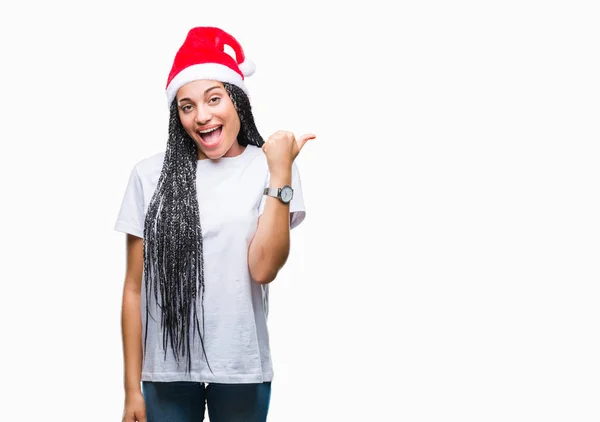年轻的编织头发非洲裔美国女孩戴着圣诞帽在孤立的背景微笑着与快乐的脸看 用拇指指着一边 — 图库照片