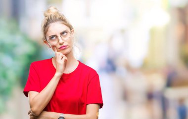 Kırmızı t-shirt ve gözlük üzerinde giyen genç güzel sarışın kadın arka plan çene düşünme hakkında soru, dalgın ifade üzerine el ile izole. Düşünceli yüzüyle gülümsüyor. Şüphe kavramı.