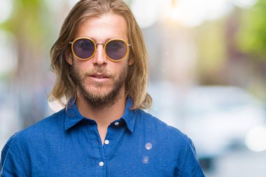 Genç yakışıklı adam uzun saçlı yüzünde ciddi bir ifade ile izole arka plan üzerinde güneş gözlüğü takıyor. Basit ve doğal kameraya bakıyor.