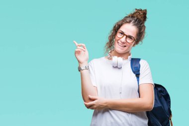 Yüzünde kocaman bir gülümseme ile izole arka plan üzerinde sırt çantası ve kulaklıklar giyen genç esmer öğrenci kız kameraya bakarak yan el ve parmak ile işaret.