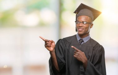 Genç Afro-Amerikan erkek gülümseyen ve iki elleri ve parmakları ile tarafına işaret kamera bakarak izole arka plan üzerinde mezun oldu.