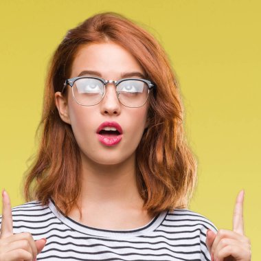 İzole arka plan gözlüklü genç güzel kadın hayrete ve yukarıya bakıyor ve parmakları ile işaret ve silah kaldırdı şaşırttı.