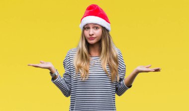 İzole arka plan clueless ve şaşkın bir ifadeyle kollar ve eller kaldırdı üzerinde Noel şapka giyen genç güzel sarışın kadın. Şüphe kavramı.