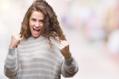 Gülümseyen ve başarı için çığlık izole arka plan çok mutlu ve heyecanlı kaldırdı, silah ile kazanan jest yapıyor üzerinde kış kazak giyen güzel esmer kıvırcık saçlı Genç kız. Kutlama kavramı.