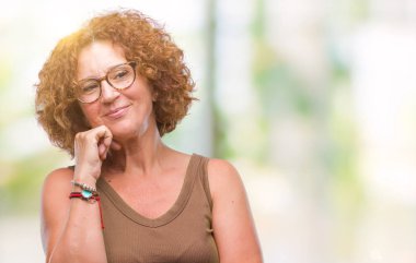 Orta yaş İspanyol kadın gözlük ile izole arka plan üzerinde çene düşünme hakkında soru, dalgın ifade ver. Düşünceli yüzüyle gülümsüyor. Şüphe kavramı.