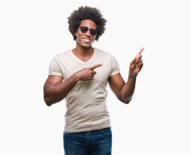 Gülümseyen ve iki elleri ve parmakları ile tarafına işaret kamera bakarak izole arka plan üzerinde güneş gözlüğü takıyor Afro Amerikalı adam.