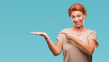 Atrractive üst düzey beyaz Kızıl saçlı kadın hayrete ve el ile sunulması ve parmak ile işaret ederken kameraya gülümseyen izole arka plan.