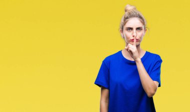 Dudaklarında parmak ile sessiz olmanı izole arka plan üzerinde mavi t-shirt giyen genç güzel sarışın ve mavi gözlü kadın. Sessizlik ve gizli kavramı.