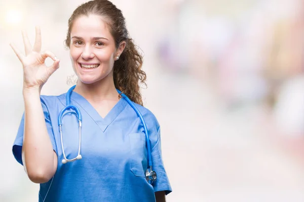 Young brunette doctor girl wearing nurse or surgeon uniform over isolated background smiling positive doing ok sign with hand and fingers. Successful expression.