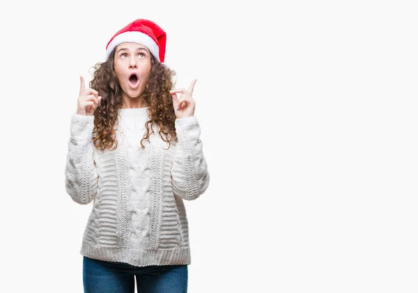 年轻的黑发女孩在孤立的背景下戴着圣诞帽 惊讶和惊讶地抬头 用手指指指点点 举起双臂 — 图库照片
