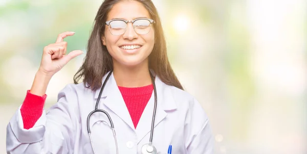 Young arab doctor woman over isolated background smiling and confident gesturing with hand doing size sign with fingers while looking and the camera. Measure concept.