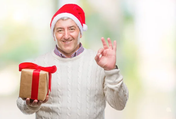 英俊的老人戴着圣诞帽子和持有礼物在孤立的背景做 符号与手指 优秀的符号 — 图库照片