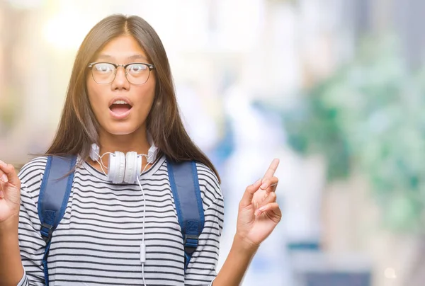 年轻的亚洲学生妇女戴着耳机和背包在孤立的背景下惊讶和惊讶地看着手指和举起手臂 — 图库照片