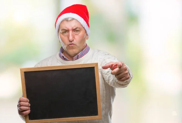 ハンサムな年配の男性の身に着けているクリスマスの帽子とカメラと指で指している孤立した背景の上持株黒板 手に署名 正面からポジティブで自信のジェスチャー — ストック写真