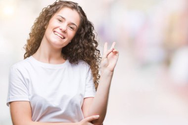 Güzel esmer kıvırcık saçlı Genç kız mutlu yüz zafer işareti yaparken kameraya göz kırpıyor ile gülümseyen izole arka plan üzerinde rahat t-shirt giyiyor. İki numaralı.