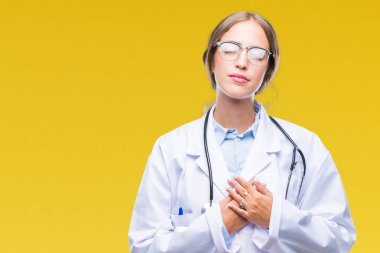 Göğüs kapalı gözlerle ellerinde ve yüzünde minnettar jest ile gülümseyen izole arka plan üzerinde tıbbi üniforma giyen güzel genç sarışın doktor kadın. Sağlık kavramı.