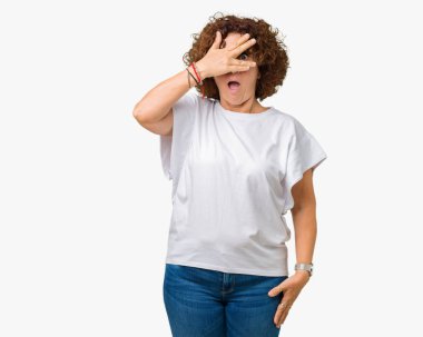 Mahcup ifadeyle parmaklarımın arasından seyir yüz ve gözler el ile kapsayan şok izole arka plan bakmak üzerinde beyaz t-shirt giyen güzel orta ager üst düzey kadın.