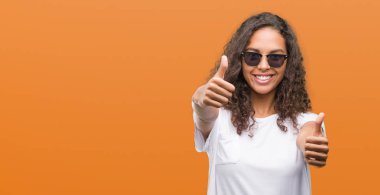 Onaylama güneş gözlüğü takmış genç İspanyol kadın el ile pozitif jest yapıyor gülümseyen ve başarı için mutlu yaşasın. Kamera, kazanan jest arıyorsunuz.