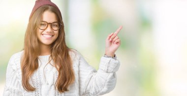 Yüzünde kocaman bir gülümseme ile izole arka plan üzerinde gözlük ve kış şapka giyen genç güzel esmer hipster kadın kameraya bakarak yan el ve parmak ile işaret.