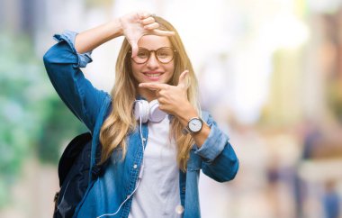 Elle yapma çerçeve ve parmakları ile mutlu yüz gülümseyen izole arka plan üzerinde kulaklık ve gözlük giyen genç güzel sarışın öğrenci kadın. Yaratıcılık ve Fotoğrafçılık kavramı.