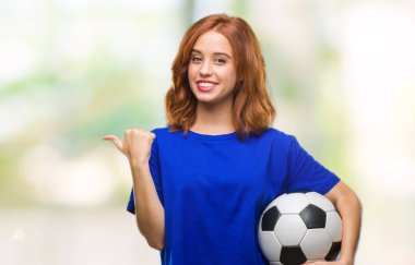 İzole arka plan işaret ve başparmak kadar mutlu yüz gülümseyen tarafı ile gösterilen futbol futbol topu tutan genç güzel kadın